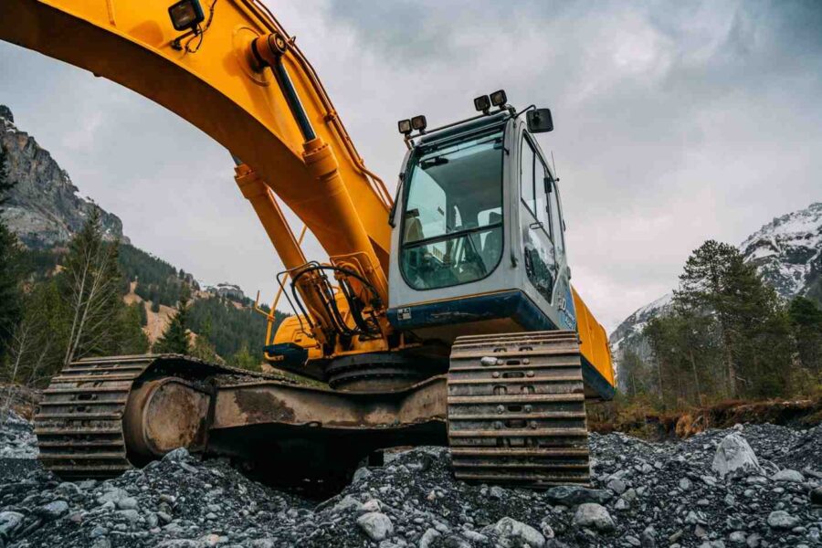 Low-angle view of a yellow tracked excavator perched on rocky terrain with forested mountains and cloudy sky in the background.