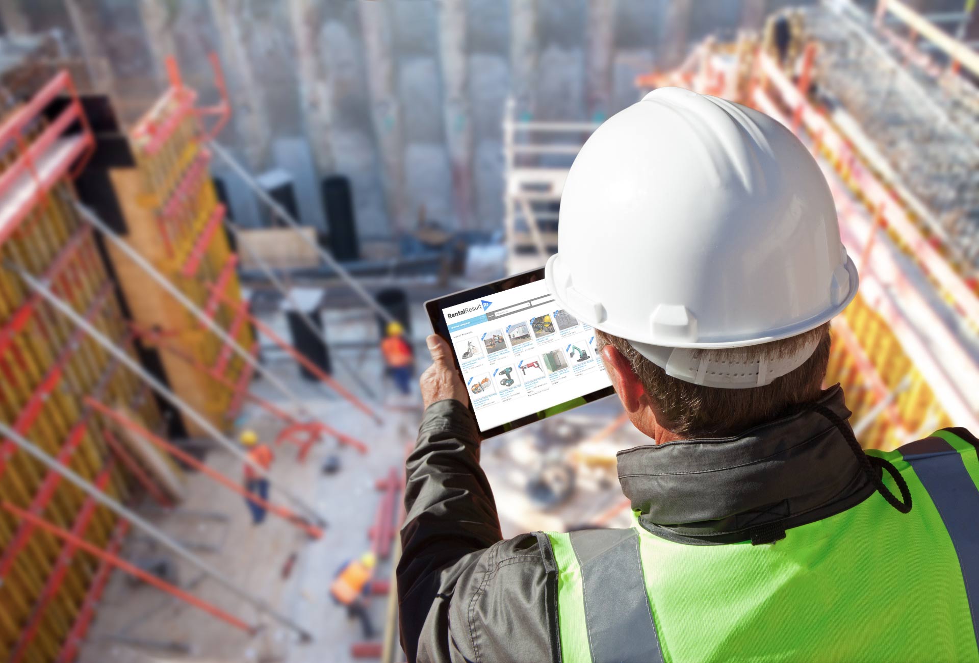 Construction supervisor in a white hard hat and high-vis vest holding a tablet showing equipment listings, overlooking a blurred busy building site with workers and scaffolding.