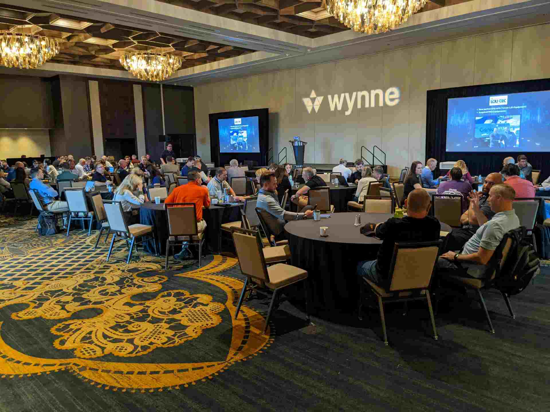 People seated at round tables in a conference room with chandeliers, attending an event.