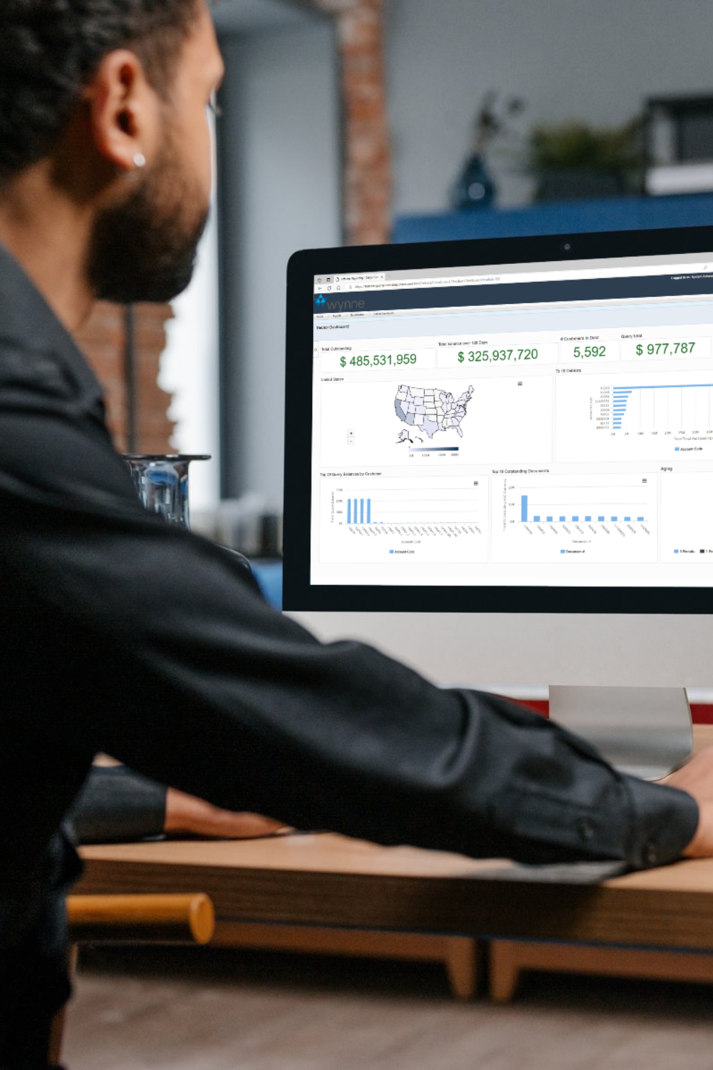 Person seated at a desk viewing a Wynne ERP Reporter business intelligence dashboard on a desktop screen, showing key financial KPIs, a US map visualization, and multiple bar charts.